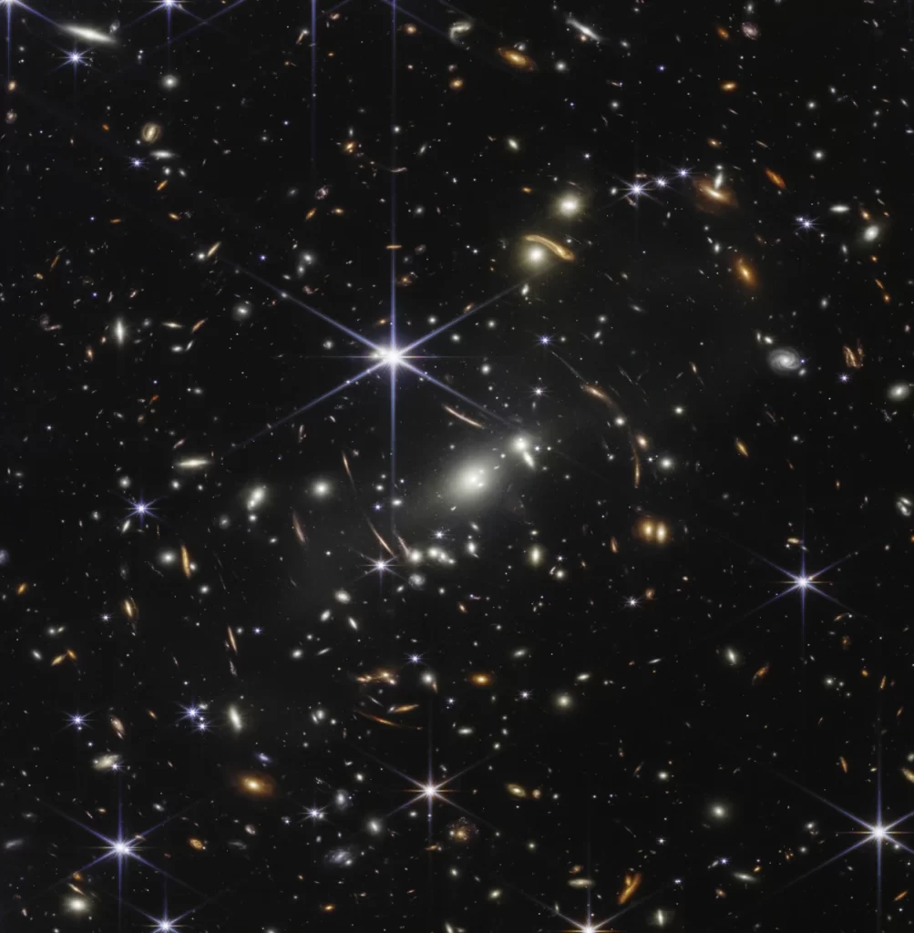 La prima immagine di James Webb è un Deep Field. Per quanto possa sembrare strano, la porzione di cosmo raccolta in questa immagine è paragonabile alla grandezza di un granello di sabbia posto ad 1m dai nostri occhi rispetto all'intera volta celeste. Al centro dell'immagine l'ammasso di galassie SMACS 0723 che genera la lente gravitazionale.